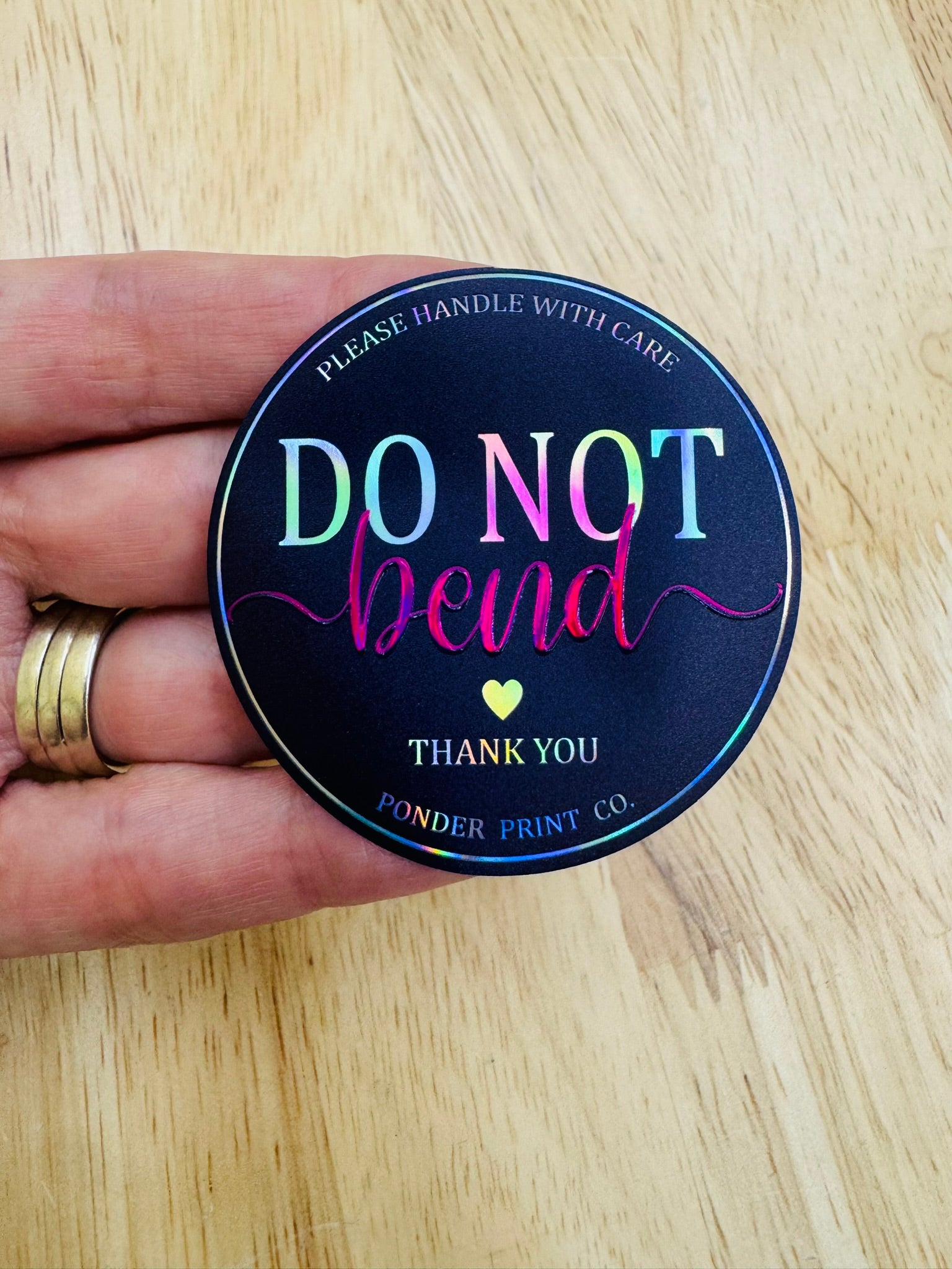 A round holographic sticker held by a hand, featuring holographic rainbow text that reads 'DO NOT bend' and 'THANK YOU' with 'Ponder Print Co.' at the bottom, set against a light wood grain background.