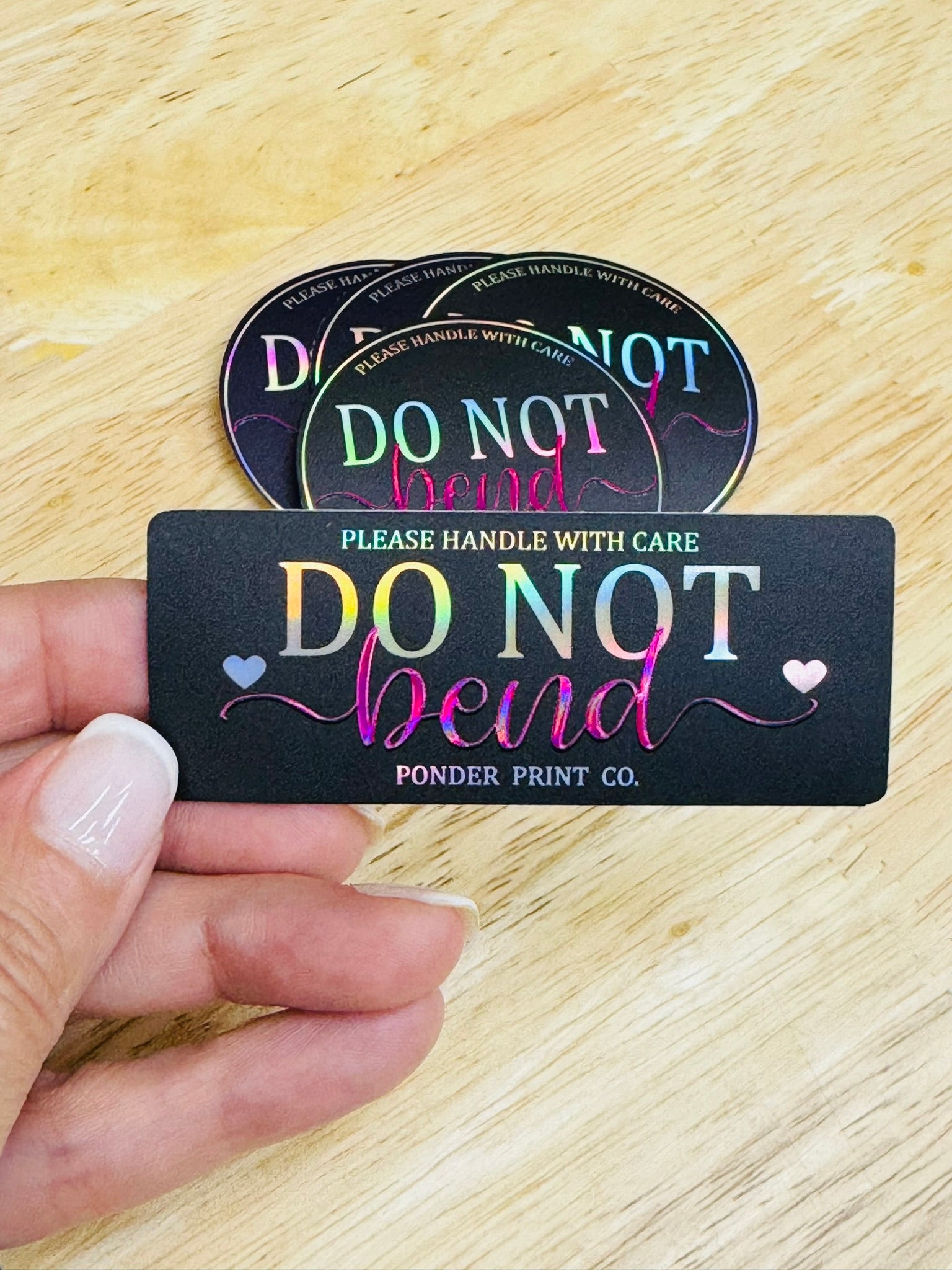 A hand holds a rectangular black sticker that reads 'DO NOT bend' in iridescent lettering, with two round matching stickers stacked behind it on a light wood surface.