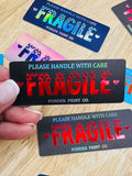 A hand holds a black rectangular sticker with the words 'PLEASE HANDLE WITH CARE' and 'FRAGILE' in red, with 'FRAGILE' in a distressed red font. Other colorful stickers are scattered on a light wood surface in the background.