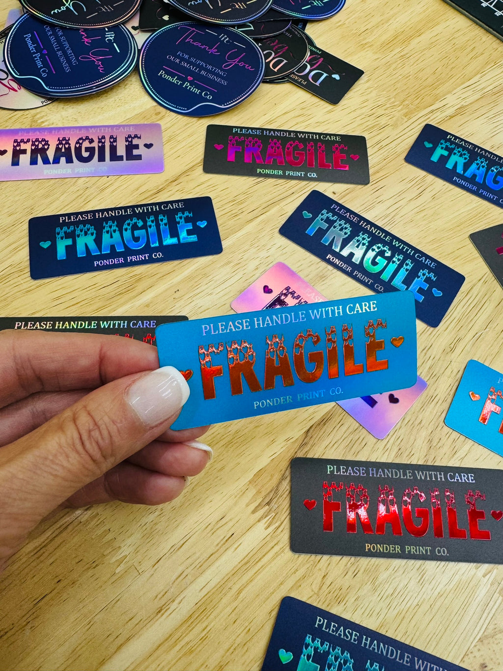 A hand holds a bright blue rectangular sticker with 'FRAGILE' in red and white, surrounded by other colorful 'FRAGILE' stickers on a light wood tabletop.