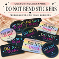 A set of custom holographic 'DO NOT BEND' stickers displayed on a light beige fabric background. The stickers feature a black base with a vibrant iridescent rainbow holographic foil effect, with text reading 'DO NOT BEND', 'PLEASE HANDLE WITH CARE', and 'THANK YOU' in pink, blue, and purple.