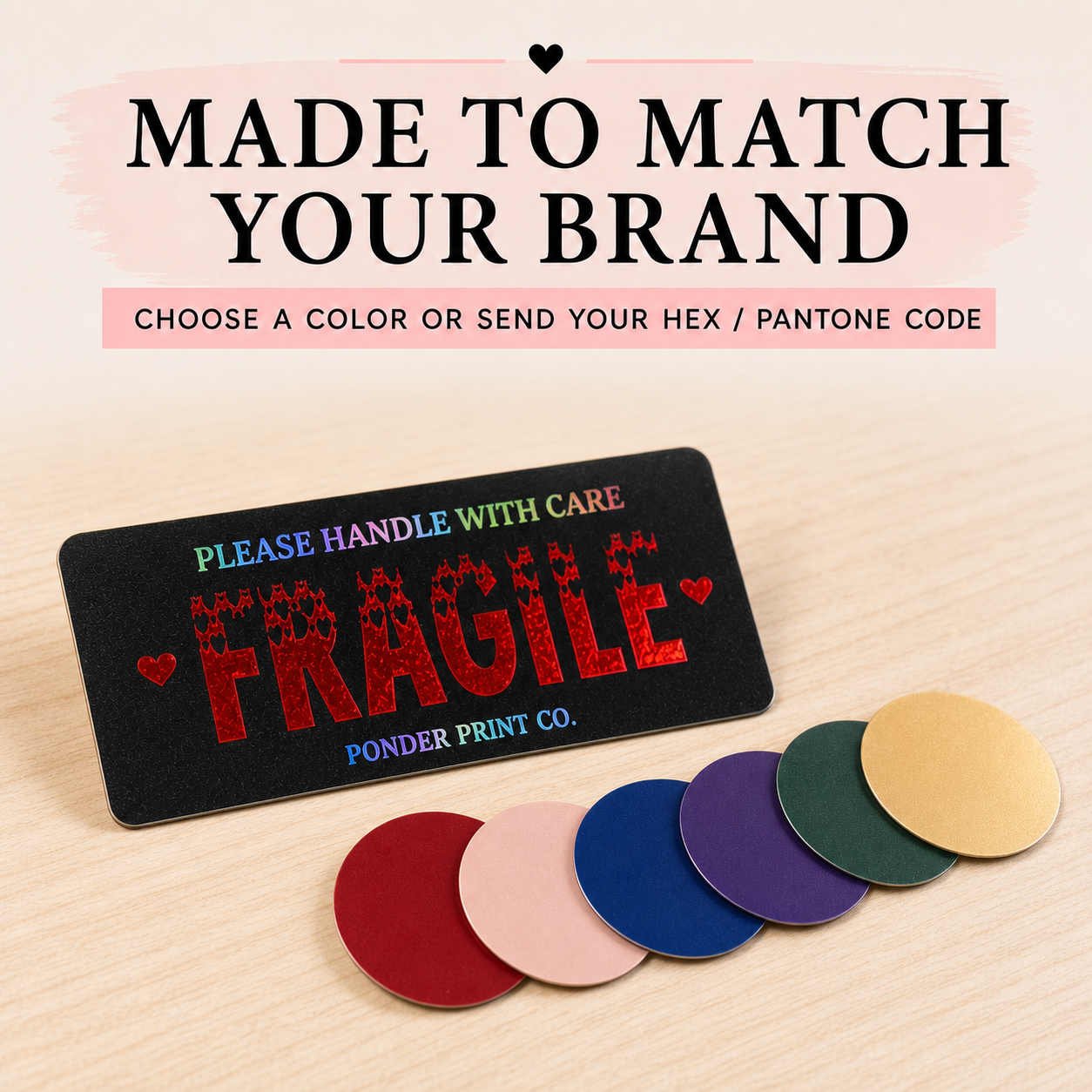 A black rectangular Fragile Sticker  with colorful text reading 'PLEASE HANDLE WITH CARE' and large red glitter-style 'FRAGILE' text, shown on a light wood surface with five circular color swatches below. Text at the top says 'MADE TO MATCH YOUR BRAND' and 'CHOOSE A COLOR OR SEND YOUR HEX / PANTONE CODE.
